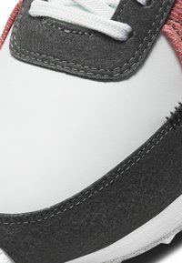 Gros plan sur l'avant de la basket montrant une tige en cuir blanc, des empiècements en daim noir, des accents en tissu rouge, et des lacets blancs passés dans des œillets en daim noir.