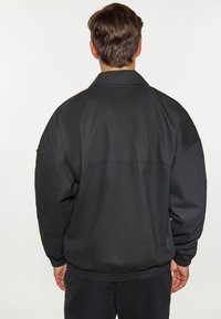 Jachetă bomber neagră cu o croială liberă, manșete și tiv elastic, și o textură mată netedă. Prezintă un guler plat și un design minimalist.