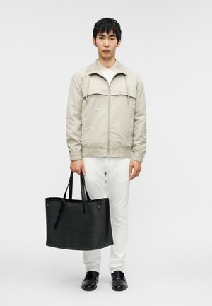 Veste beige à fermeture éclair avec capuche, chemise blanche en dessous, pantalon clair, sac cabas noir structuré, et chaussures noires avec boucles.