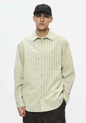 Jeune homme portant une chemise boutonnée rayée vert clair et une casquette de baseball noire, debout devant un fond blanc uni.