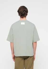 Άνδρας με κοντά σγουρά μαλλιά, φορά χαλαρό ανοιχτό γκρι κοντομάνικο πουκάμισο με λευκή ετικέτα στο άνω μέρος της πλάτης και χακί παντελόνι, στραμμένος προς τα πίσω.