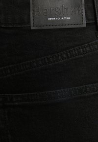 Jeans de mezclilla negra con una superficie texturizada, que presentan un parche de cuero etiquetado con "Colección de Denim Bershka" y detalles de costura contrastantes.
