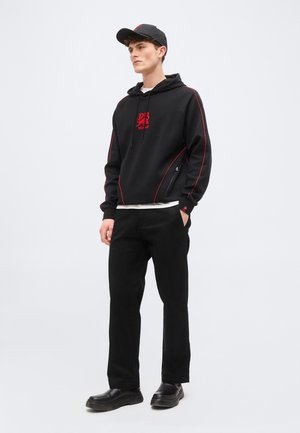 Noor mees, kes kannab musta kapuutsiga pusa, kus on punane logo ja õmblused, mustad püksid, must nokamüts ja mustad jalanõud, seisab halli ühtlase tausta ees.