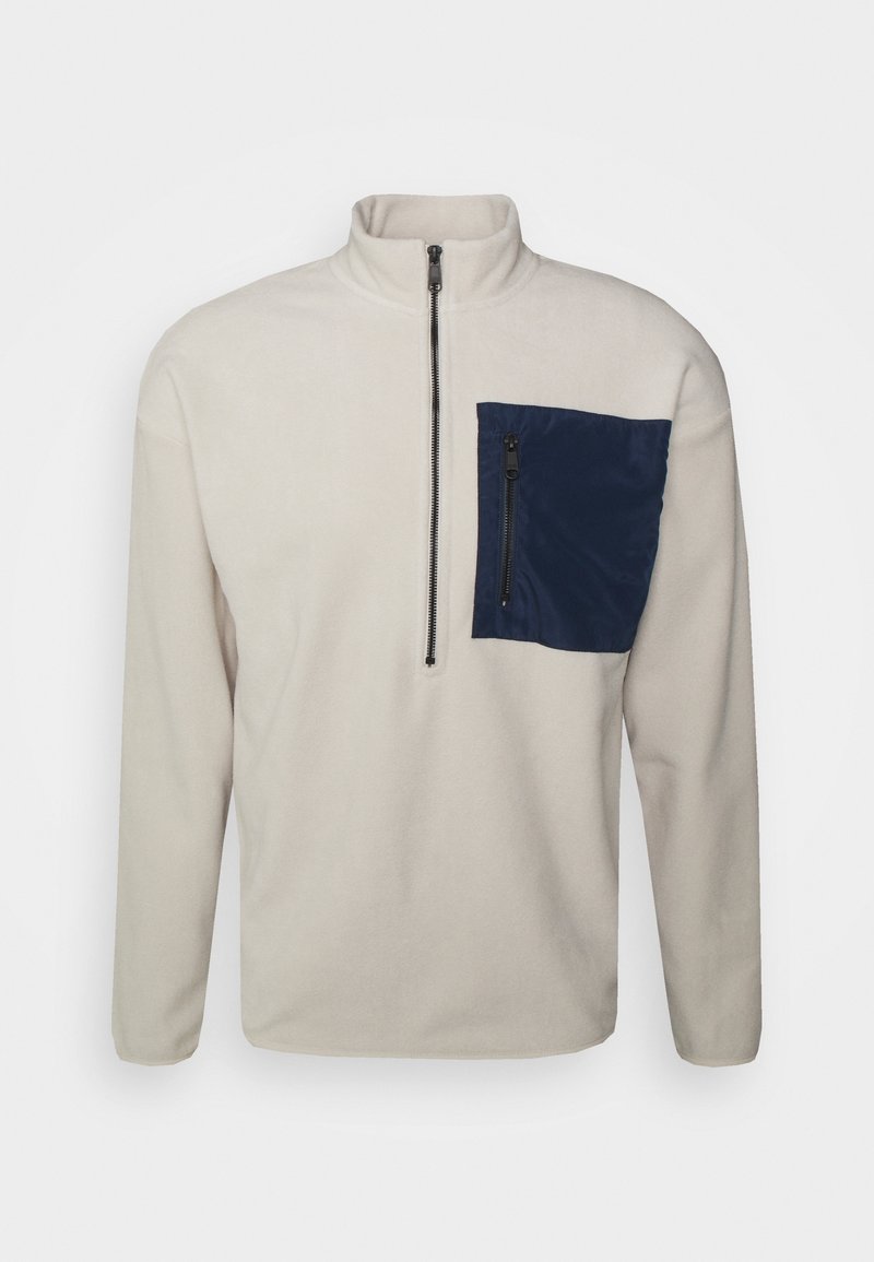 Lys beige fleece-pullover med en kvart lynlås og en stor marineblå lynlåslomme på venstre side af brystet.