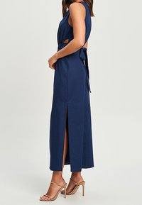 Granatowa sukienka bez rękawów z wycięciami po bokach, wiązanym w pasie oraz dwoma rozcięciami z przodu; wykonana z gładkiego materiału o długim, zwiewnym kroju.