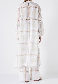 Lange weiße bestickte Tunika mit floralem Muster und Ausschnittdetails. Weicher Stoff, lange Ärmel und lockere Passform. Kombiniert mit weißen Hosen.