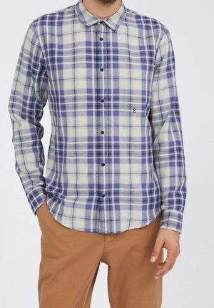 Mann trägt ein langärmeliges Hemd mit blau-weißem Karomuster und helle braune Hose, steht mit einer Hand in der Tasche.