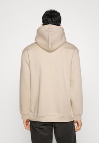 Felpa beige con cappuccio e vestibilità rilassata, caratterizzata da un ampio cappuccio e polsini a coste. Tessuto morbido, senza motivi o loghi visibili.
