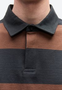 Κοντινό πλάνο ενός ατόμου που φοράει καφέ και μαύρη ριγέ πόλο μπλούζα με μαύρο γιακά και ένα κουμπί κουμπωμένο.