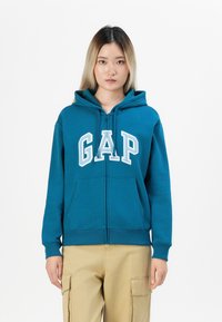 Teal zip-up hoodie with front pockets and white "GAP" lettering. Features a drawstring hood and ribbed cuffs at sleeves and hem.