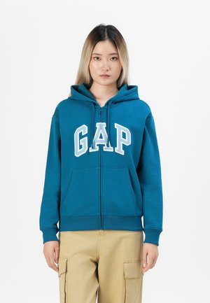 Teal hettegenser med glidelås, frontlommer og hvit "GAP"-skrift. Har snor i hetta og ribbestrikkede mansjetter på ermene og i kanten.