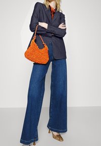 Oranssi kudottu käsilaukku, jossa on teksturoidu pintaa, yhdistetty tumman bleiserin ja sinisten leveälahkeisten farkkujen kanssa. Korkokengät, joissa on käärmeen nahkakuosinen kuvio.