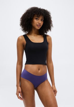 Γυναίκα με κατσαρά μαλλιά που φοράει μαύρο αμάνικο crop top και μοβ αόρατο εσώρουχο, στέκεται μπροστά σε λευκό απλό φόντο.