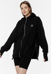 Czarny oversize hoodie z zamkiem, bocznymi kieszeniami i okrągłym logo. Posiada długie rękawy i kaptur ze sznurkiem. Miękka tekstura materiału.
