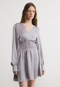 Νεαρή γυναίκα που φοράει φόρεμα από σατέν ανοιχτό μοβ χρώμα με μακριά μανίκια, βαθύ ντεκολτέ σε σχήμα V και ζώνη που συγκεντρώνει στη μέση, στέκεται μπροστά σε απλό λευκό φόντο.