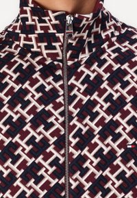 Chaqueta con cremallera que presenta un patrón geométrico en burdeos, azul marino y crema, con un cuello alto y detalle de cremallera plateada.