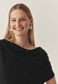 Sort off-shoulder top med en blød tekstur. Model der bærer store, skinnende, dråbeformede sølvøreringe. Neutral baggrund.