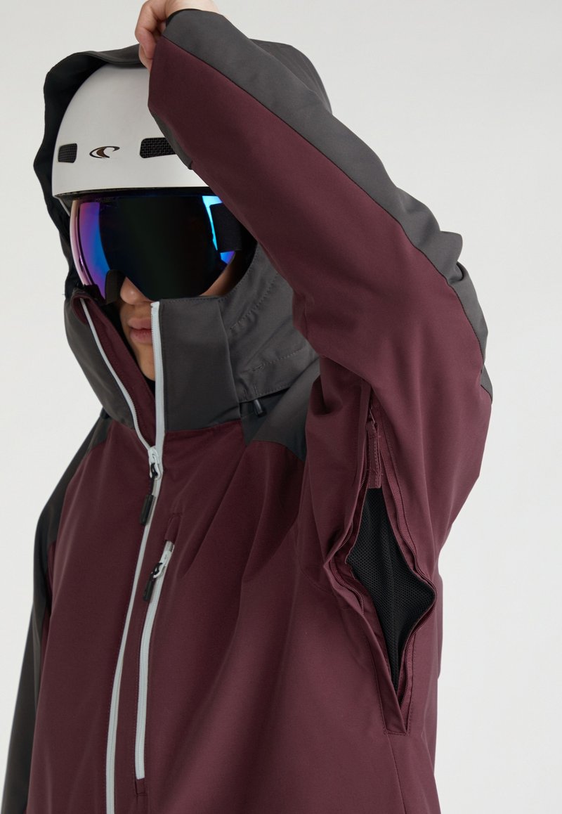 Person mit maroonem und dunkelgrauem Skijacke, die den Arm hebt und eine offene zipfelbelüftung unter dem Arm zeigt, mit weißem Helm und reflektierenden Goggles.