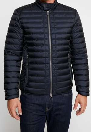 Marinblå quiltad jacka med hög krage, framzipper, två sidofickor och texturerade sidopaneler. Slät, lätt material.