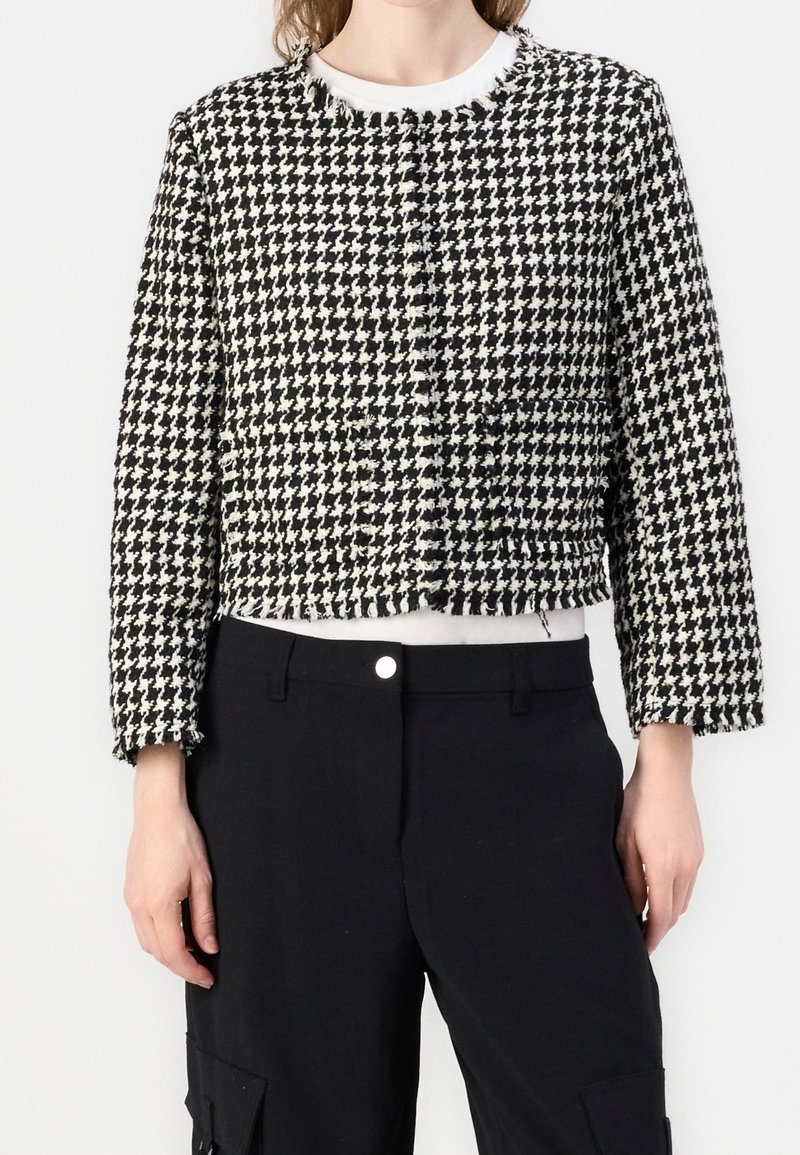 Buste féminin portant une veste courte en pied-de-poule noir et blanc sur un haut blanc, avec un pantalon noir taille haute à poches latérales.
