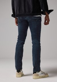 Person som bär mörkblå slim-fit jeans, svart jacka och vita sneakers med beige och svarta accenter som står framför en enfärgad bakgrund.