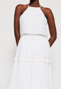 Bílá halter-style šaty s detailním krajkovým lemem na korzetu a pase, které mají splývavou sukni vyrobenou z lehkého materiálu.