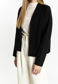 Schwarzer, offen getragener Blazer aus einem weichen Stoff mit langen Ärmeln, kombiniert mit cremefarbenen, hochtaillierten Hosen. Glattes, gepflegtes Design.