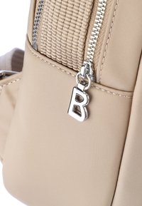 Beige ryggsäck med en texturerad tyg, silverfärgad dragkedja och en framträdande bokstav "B" som dragkedjedetalj.