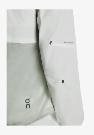 Gaiši pelēka jaka ar sieta paneli, uzrakstu “Swiss Engineering” un melnām detaļām. Materiāls šķiet viegls un gluds.