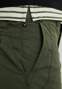Plano detalle del bolsillo de pantalones verdes con botón negro y dobladillo de camisa a rayas blanca y verde encima.