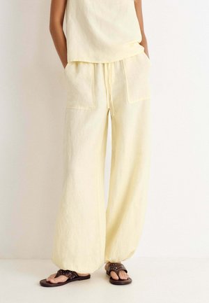 Personne portant un pantalon ample jaune clair avec de grandes poches avant et un haut sans manches assorti, associé à des sandales cloutées marron foncé.