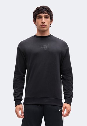 Jeune homme portant un sweat-shirt noir à manches longues avec un logo taureau subtil et un pantalon Nike noir, debout devant un fond uni.