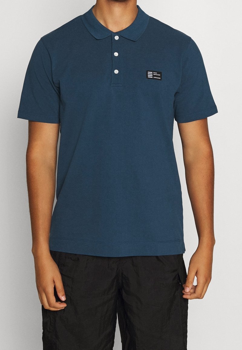 Homme portant un polo bleu foncé à manches courtes avec trois boutons blancs et un petit écusson logo noir sur la poitrine gauche.