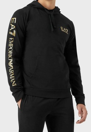 Conjunto de sudadera con capucha y pantalones negros con el logo dorado "EA7 Emporio Armani" en el pecho y la manga izquierda, llevado por una persona de pie con las manos parcialmente en los bolsillos.
