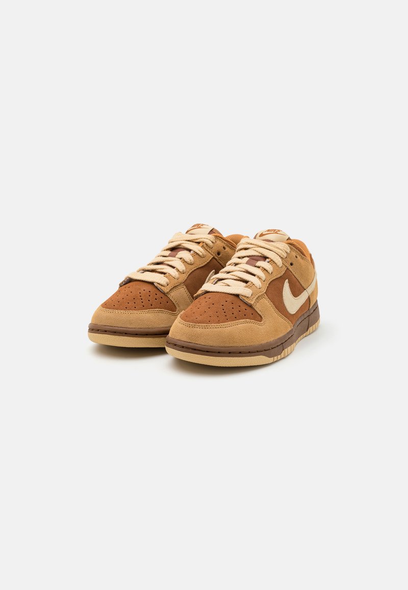 Bruine suède sneakers met een geperforeerde teenbox, contrasterende donkerbruine accenten, beige veters en een witte Nike Swoosh-logo aan de zijkant.