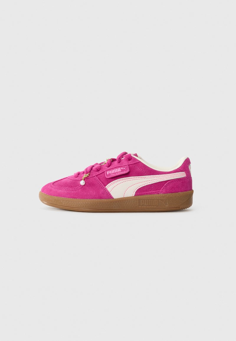 Sneaker Puma di suede rosa brillante con striscia laterale bianca, suola in gomma e perla decorativa sui lacci, su sfondo grigio chiaro.