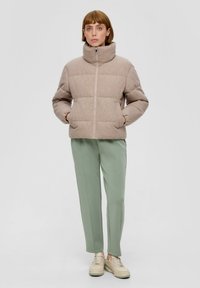 Jasnobrązowa puchowa kurtka z wysokim kołnierzem, zamkiem błyskawicznym z przodu i dwoma kieszeniami po bokach, noszona z zielonymi zwężonymi spodniami i beżowymi sneakersami.