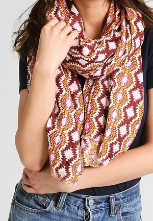 Femme portant une écharpe à motifs géométriques multicolores sur un haut noir et un jean bleu, tenant l'écharpe près de son cou.