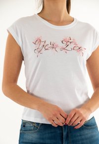 T-shirt bianca a maniche corte con ricami decorativi floreali rosa. Il tessuto è morbido e il design include accenti metallici.