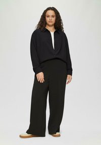 Zwarte button-up shirt met een losse pasvorm, gecombineerd met brede zwarte broek. Model draagt beige en bruine sneakers. Eenvoudig, minimalistisch ontwerp.