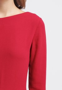 Punainen pitkähihaisen mekko, jossa on veneen kaula-aukko, sileä tekstuuri, vartalonmyötäinen siluetti ja jämäkät olkapäät, luoden modernin ilmeen.