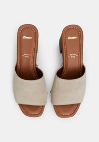 Sandales à talon bloc en suède beige avec bout ouvert et brides croisées. La semelle est en cuir marron avec embossage. Détails en surpiqûre.