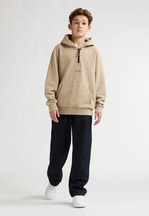 Beige hoodie met een rits bij de kraag, een zak aan de voorkant en geribde manchetten, gecombineerd met donkere broek en witte sneakers. Soepele katoenen textuur.
