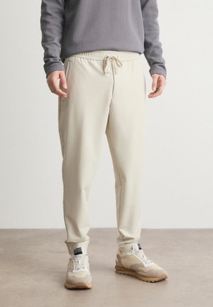Pantalon de survêtement beige avec une taille élastique et un cordon de serrage ; texture lisse ; design fuselé ; poches latérales ; associé à des baskets beiges et blanches.
