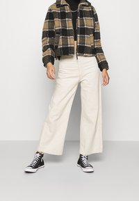 Chaqueta de cuadros recortada en negro, beige y blanco, combinada con pantalones anchos marfil y zapatillas negras de caña alta. Tejido texturizado, corte relajado.