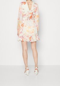 Robe florale à manches longues, dos en V, ourlet à volants et palette de couleurs claire. Portée avec des talons compensés blancs. Vue de dos affichée.