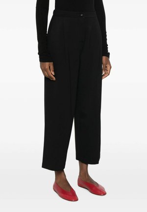 Donna che indossa pantaloni neri a gamba larga e corti, con top nero a maniche lunghe e ballerine rosse con punta, in piedi su sfondo bianco.