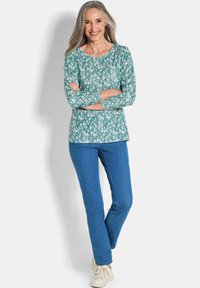Langärmliges türkises Oberteil mit geometrischem Muster, kombiniert mit blauen Jeans und hellen Sneakers. Das Model steht mit verschränkten Armen.
