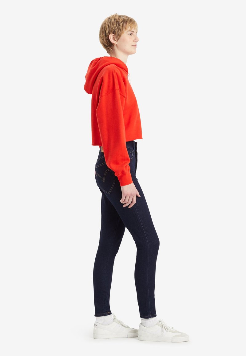Sweat à capuche rouge court avec un cordon de serrage, associé à un jean bleu foncé et des baskets blanches. La tenue présente une silhouette décontractée et ajustée.