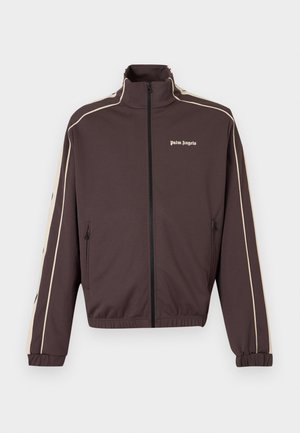 Mand iført sorte solbriller, brun zip-up jakke, hvid top, beige bukser med mørk grafisk tekst og sorte sneakers, hænderne i lommerne, kigger mod højre.